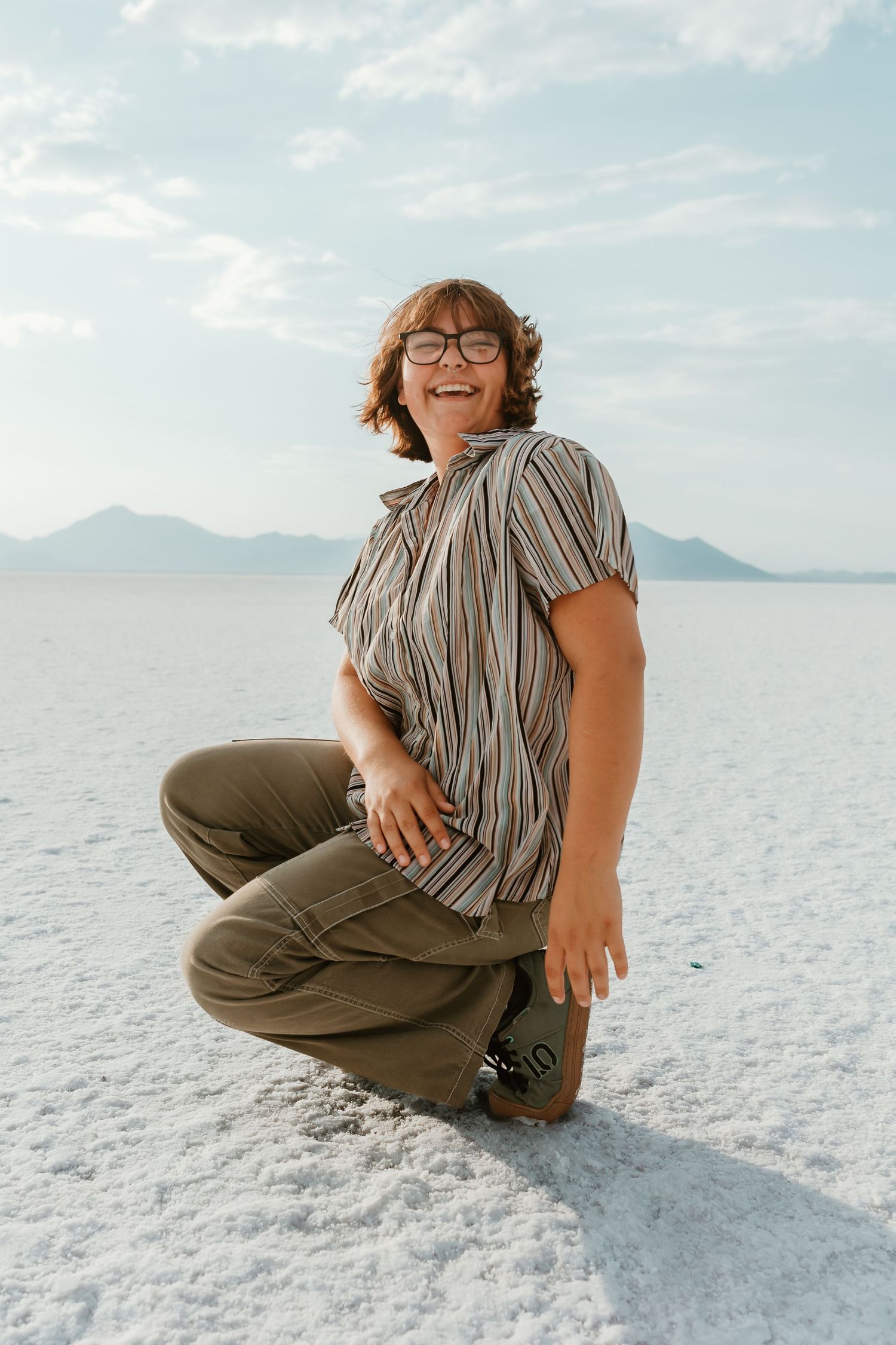 Person sitting on white salt flats with mountains in the distance wearing casual attire.