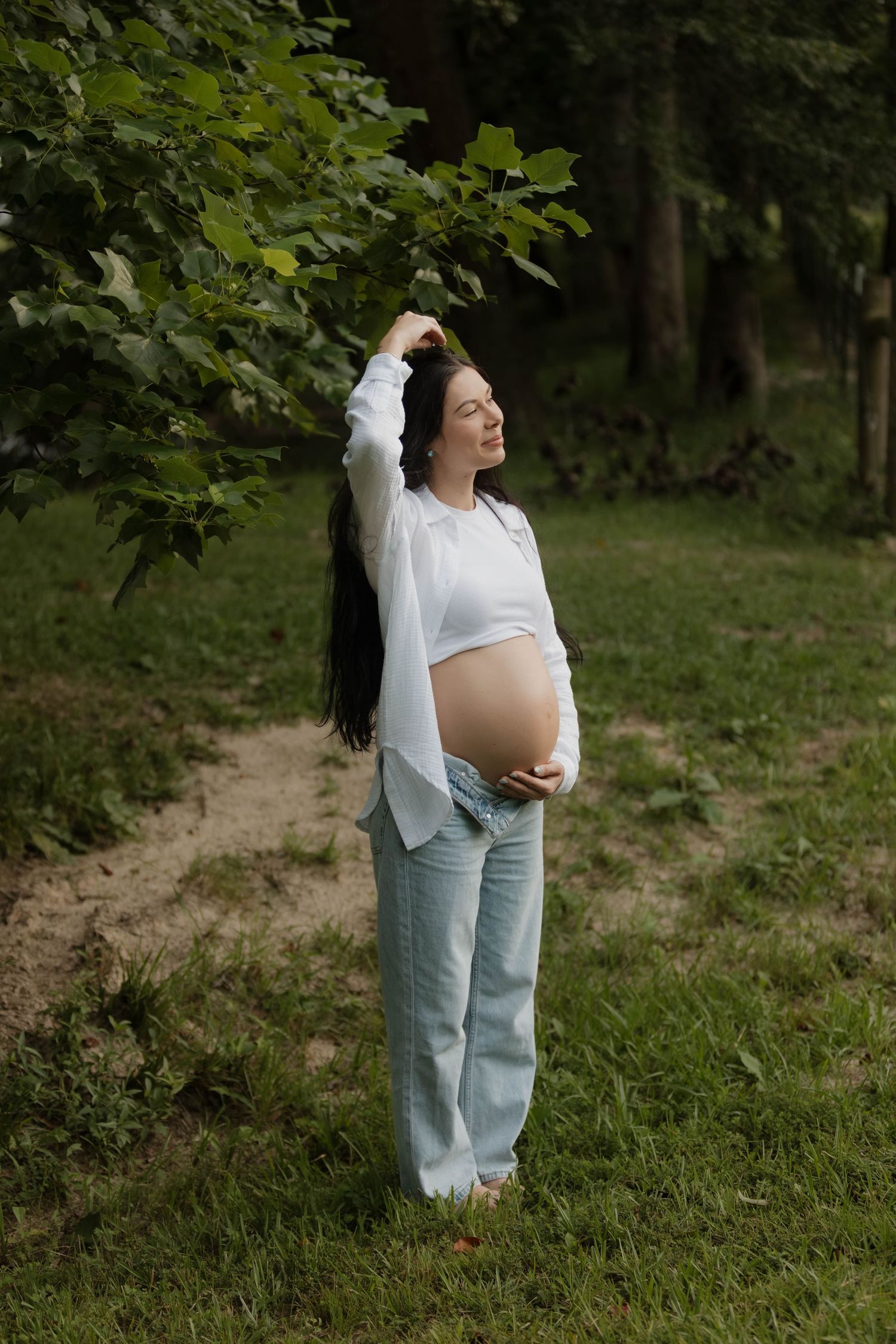 Maternity photo of pregnant woman in white top and jeans standing in lush green setting.
