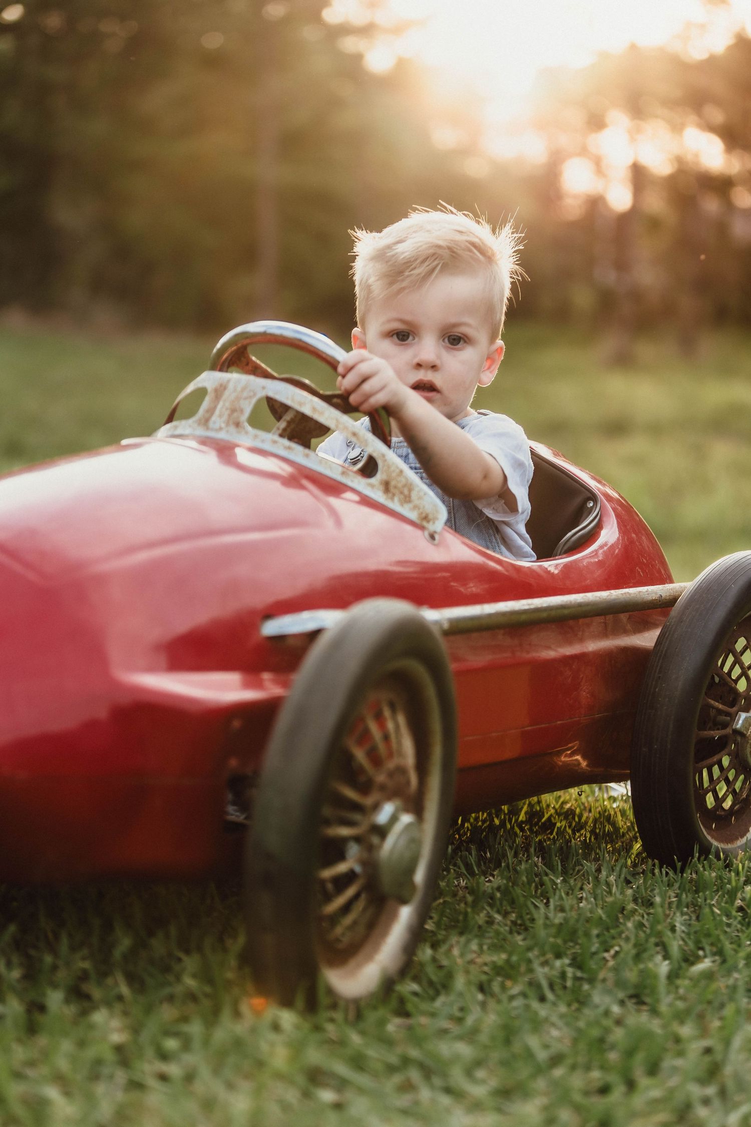 Child plays with classic red pedal car in sunlit grassy field.