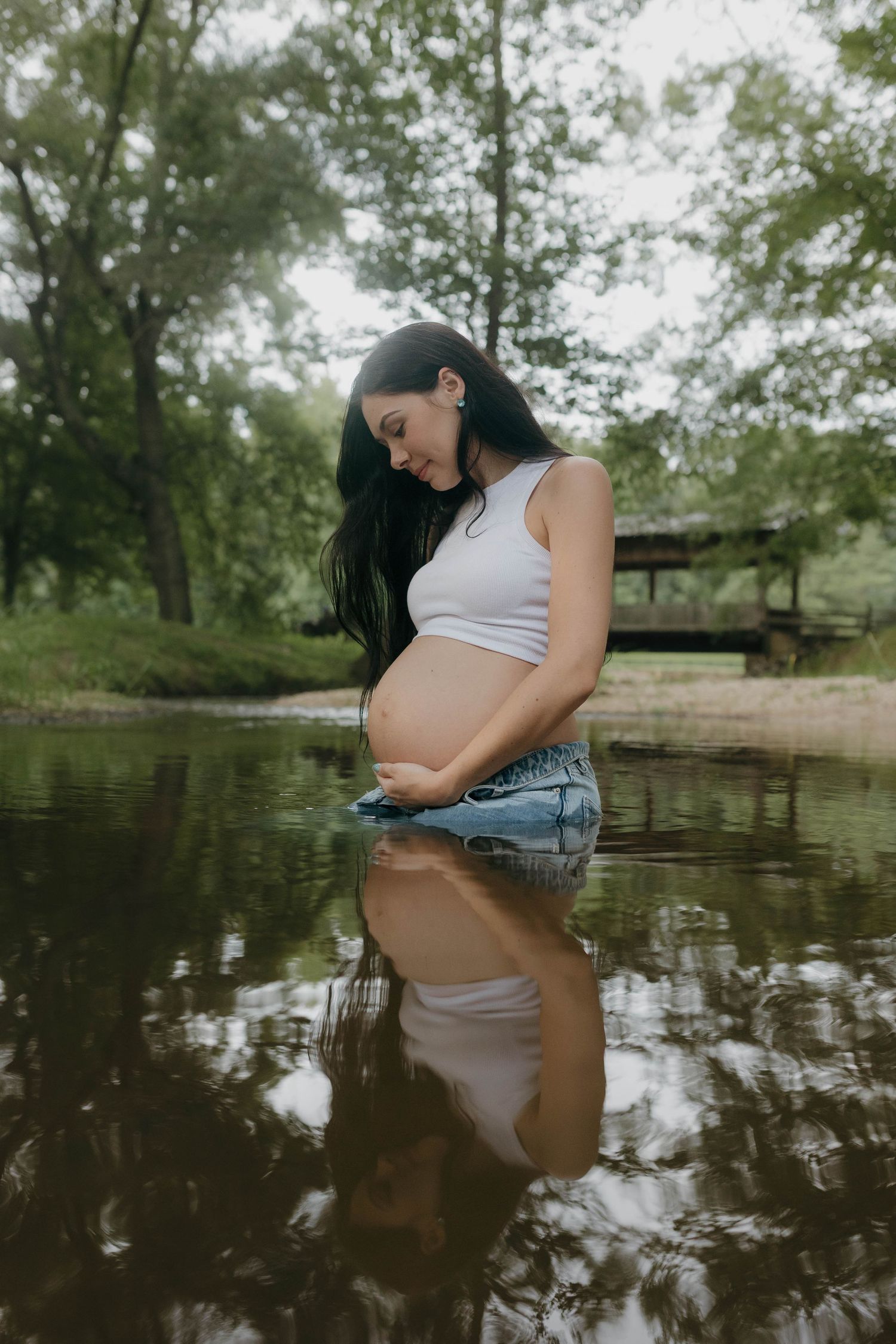 Creative maternity photo with reflection in calm water by wooded shoreline.