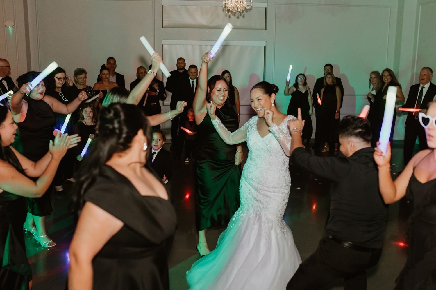Lively wedding reception dance floor with guests celebrating under chandelier lighting.