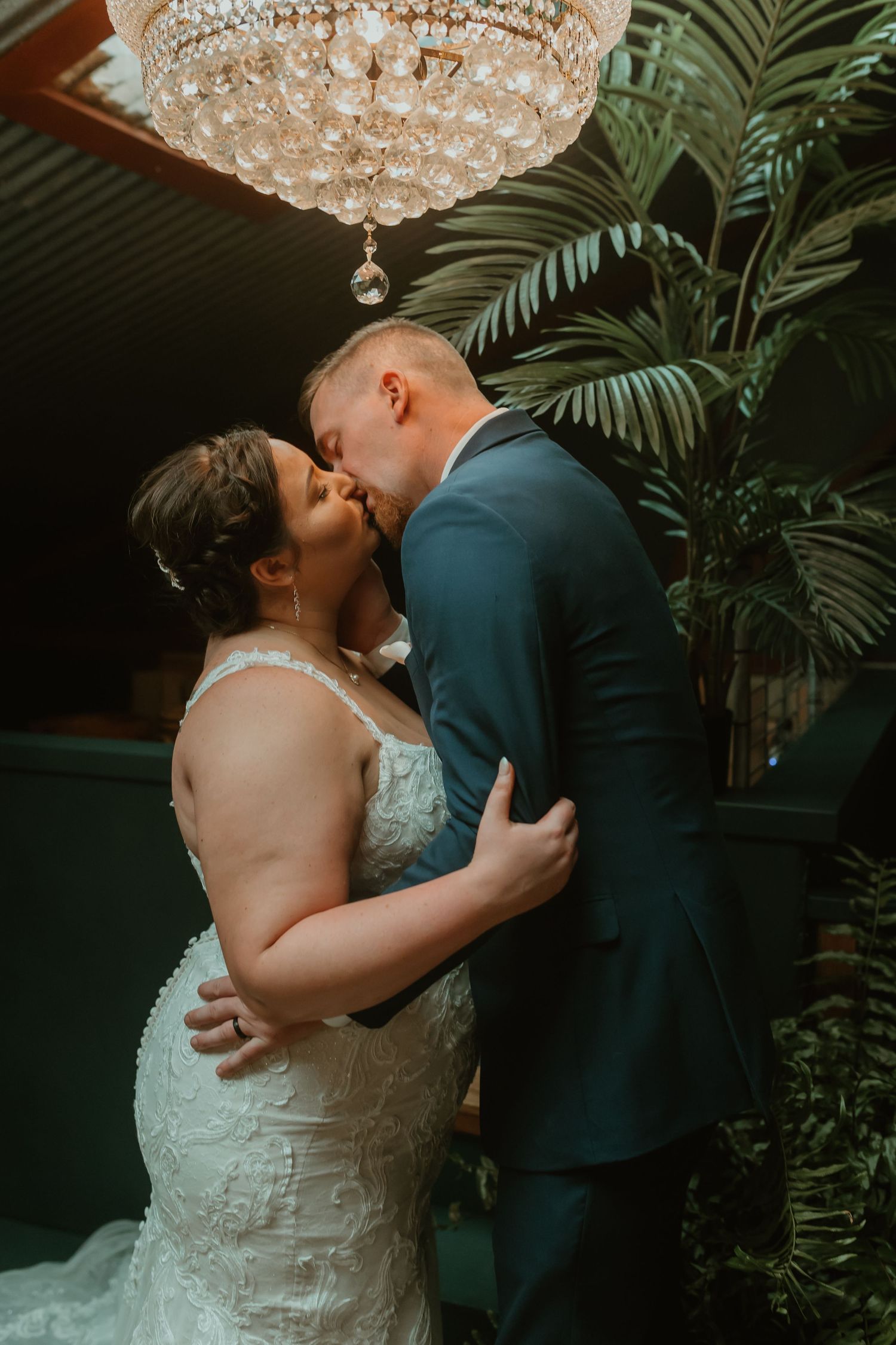 A romantic wedding moment under a crystal chandelier with lush tropical palm fronds in the background.