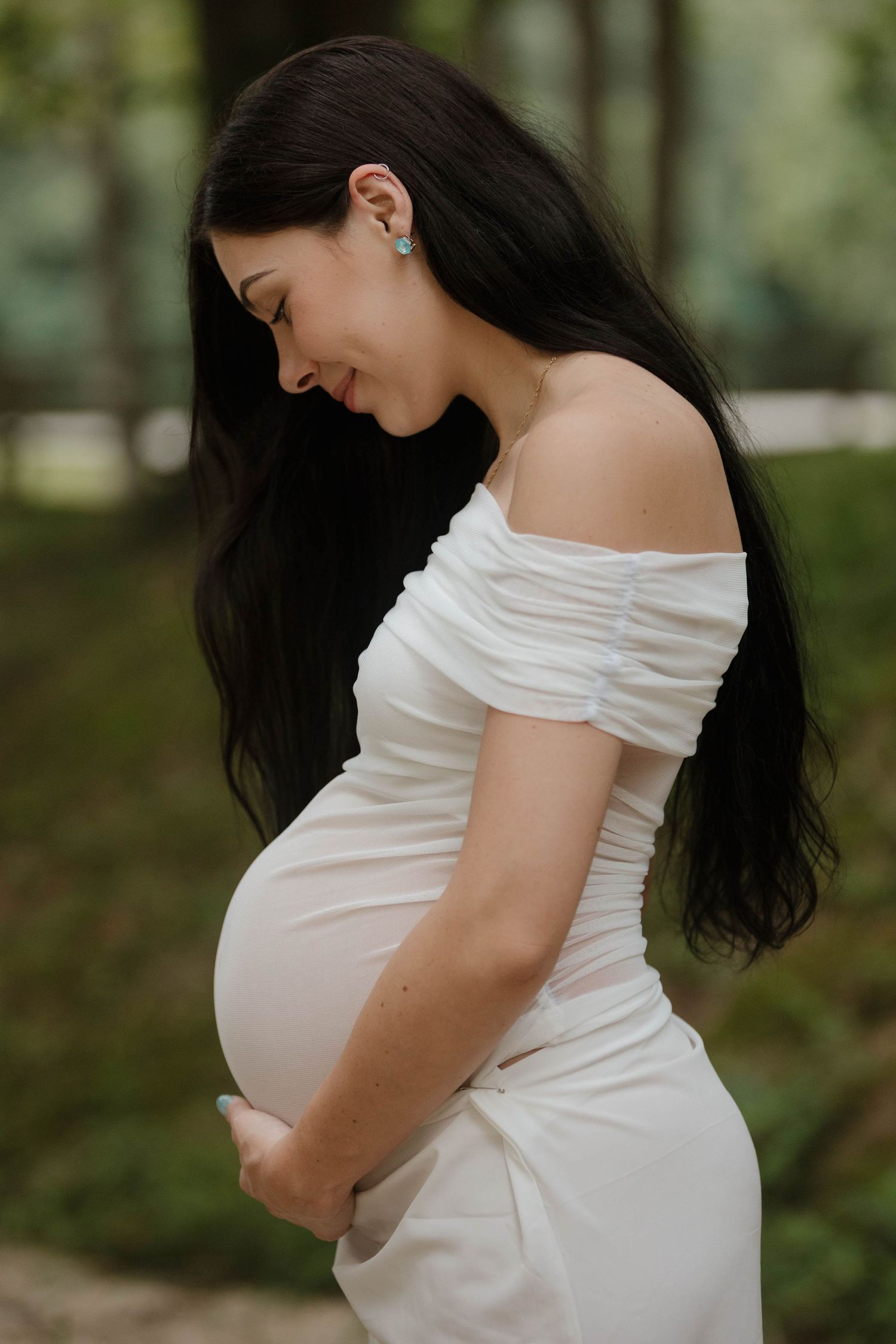 Maternity photos showing a woman in a white off-shoulder dress cradling her baby bump in a natural outdoor setting.