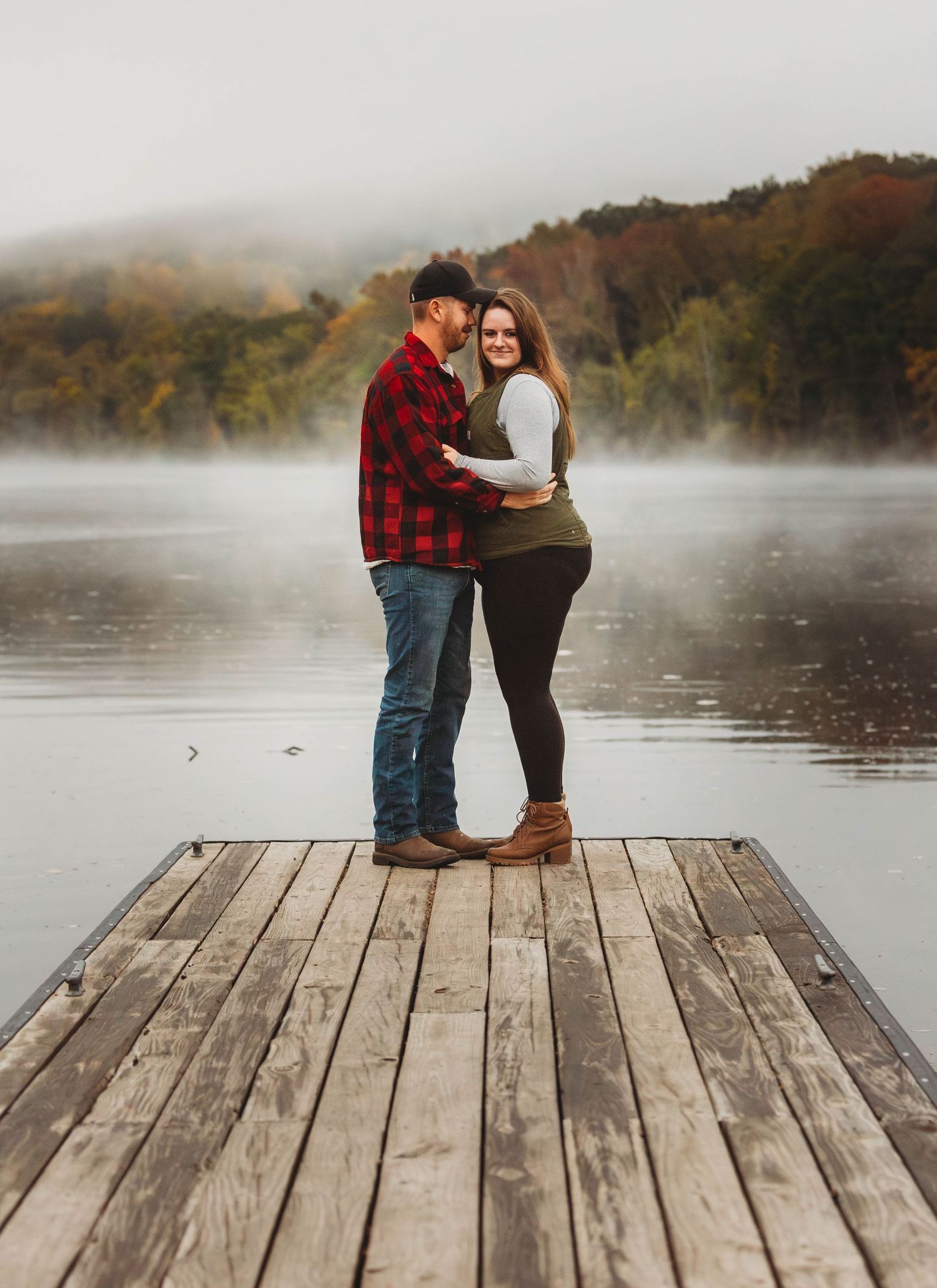 Couple embracing on wooden dock over misty lake with fall foliage in background.