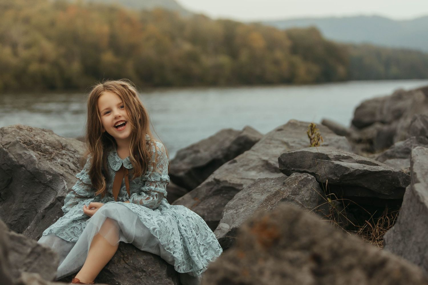 Young girl in a blue dress laughing while sitting on rocky riverbank with mountains in background.