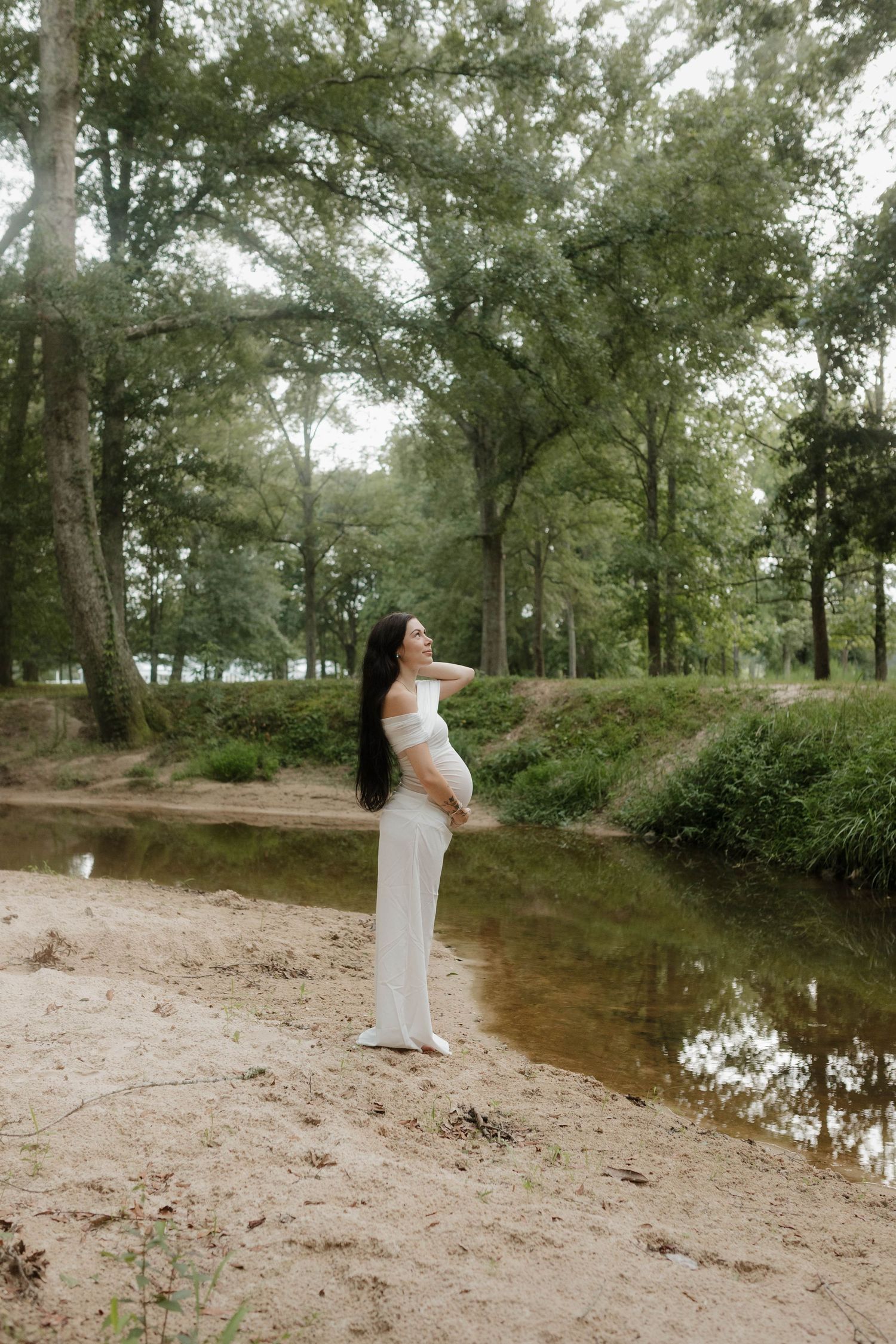 Serene maternity portrait of woman in white dress standing by riverside in wooded setting.