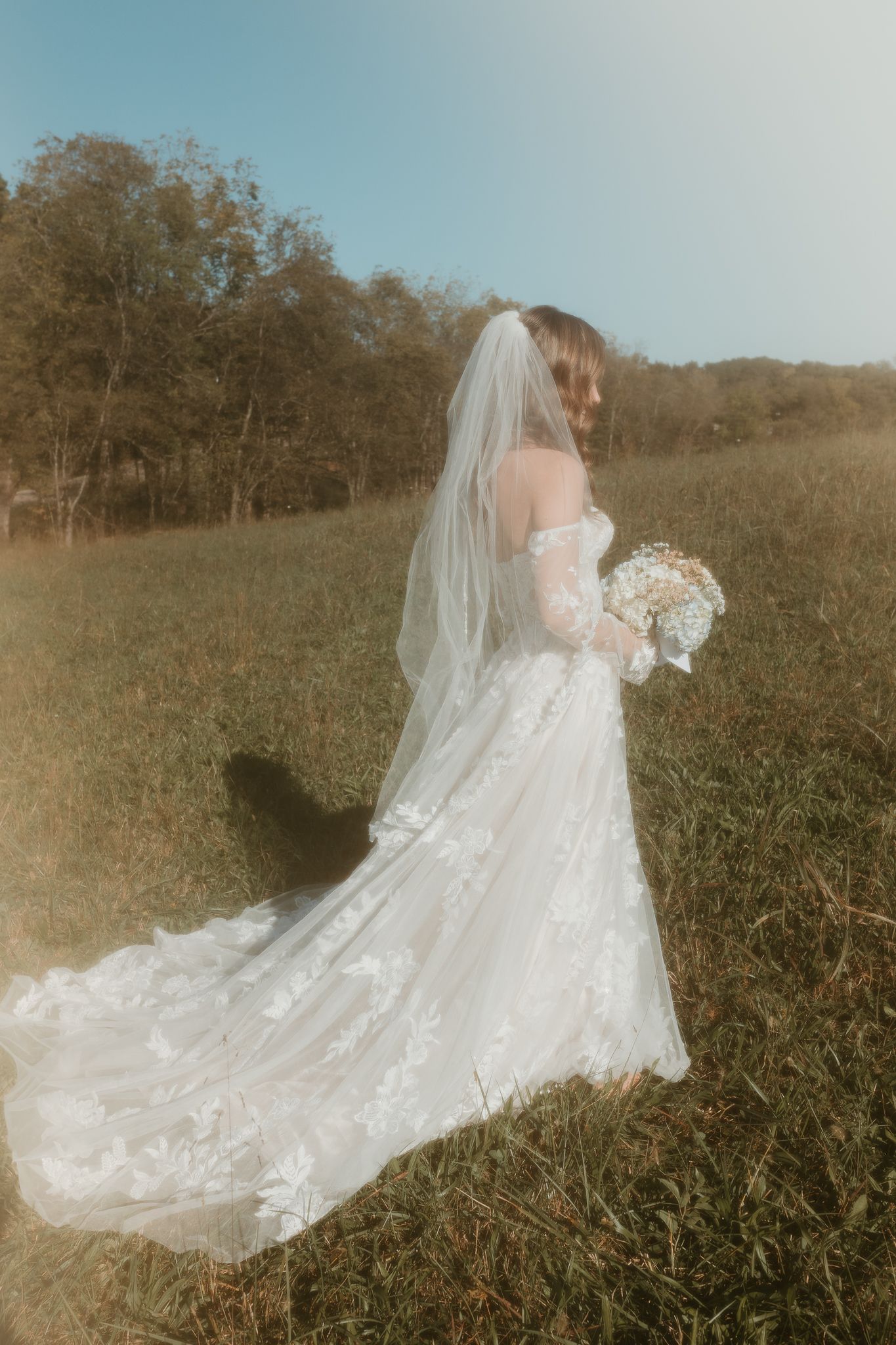 Romantic wedding dress with lace train trailing across grassy meadow at sunset.