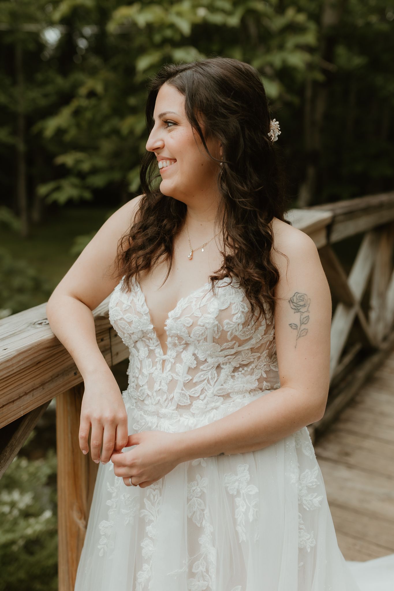 Bride in flowing lace gown poses on wooden deck surrounded by greenery.