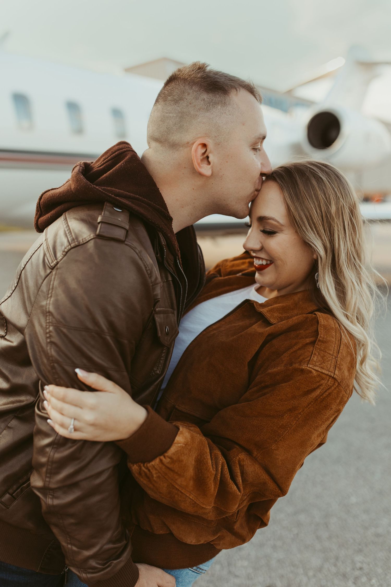 A romantic moment captured between two people wearing brown and rust colored jackets near an airplane.