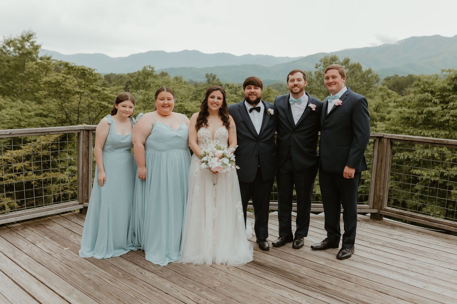 Wedding party in light blue and black attire posed on mountain overlook deck.