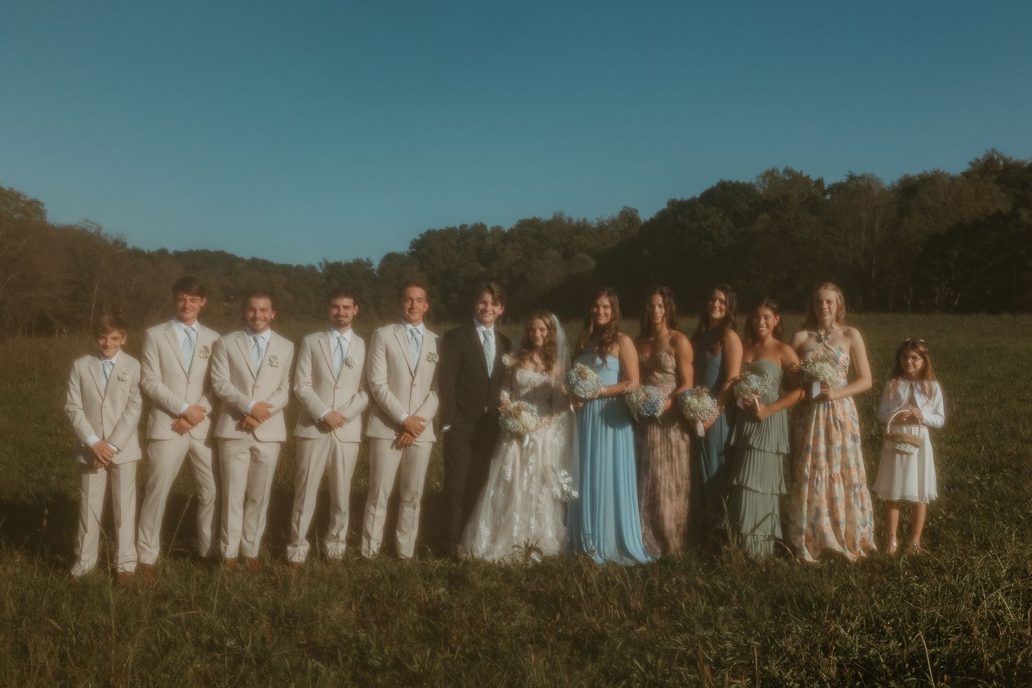 Wedding party in white and blue attire lined up for group photo in outdoor setting.