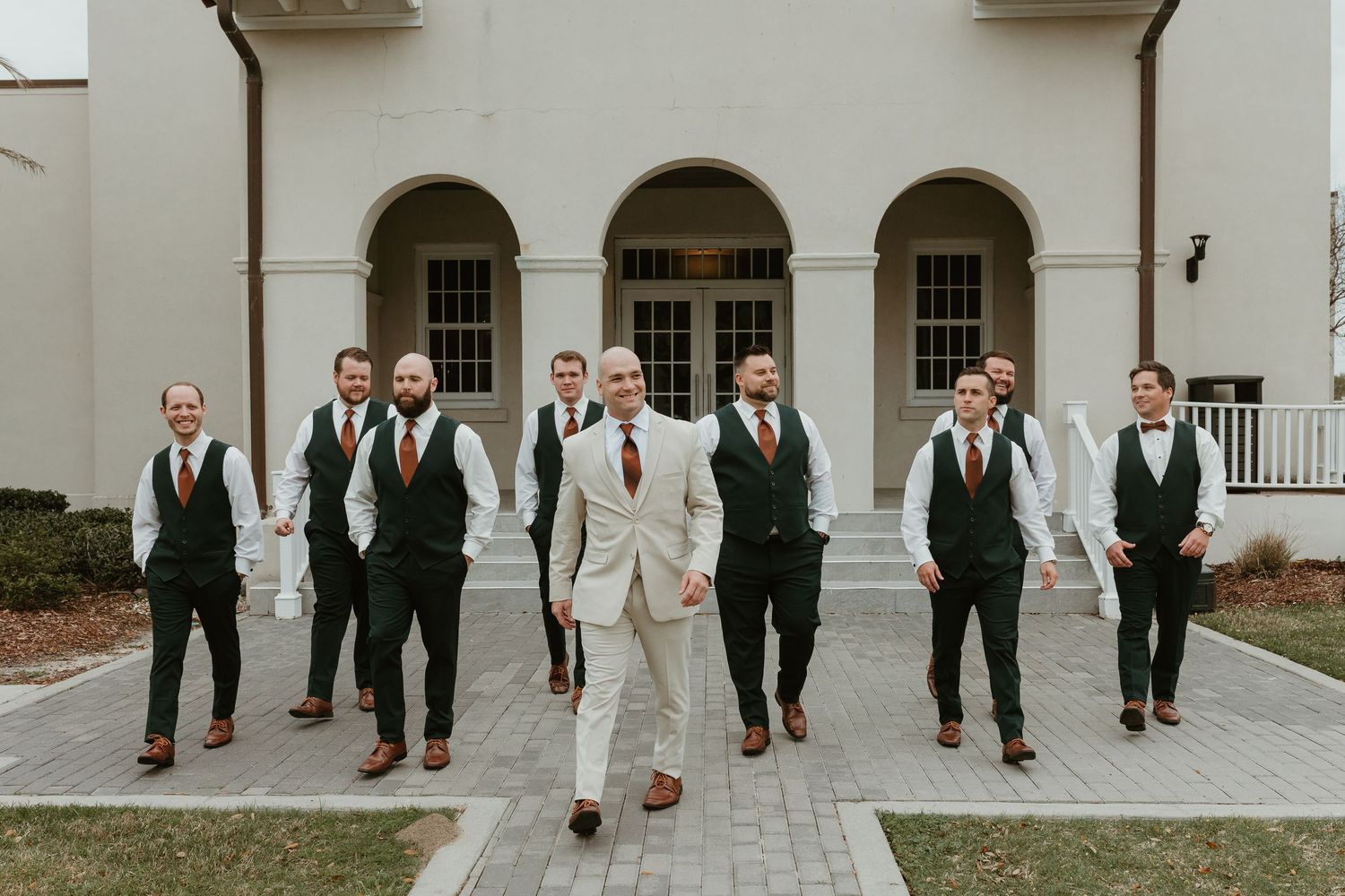 Wedding party in formal attire walking together outside a white colonial-style building.
