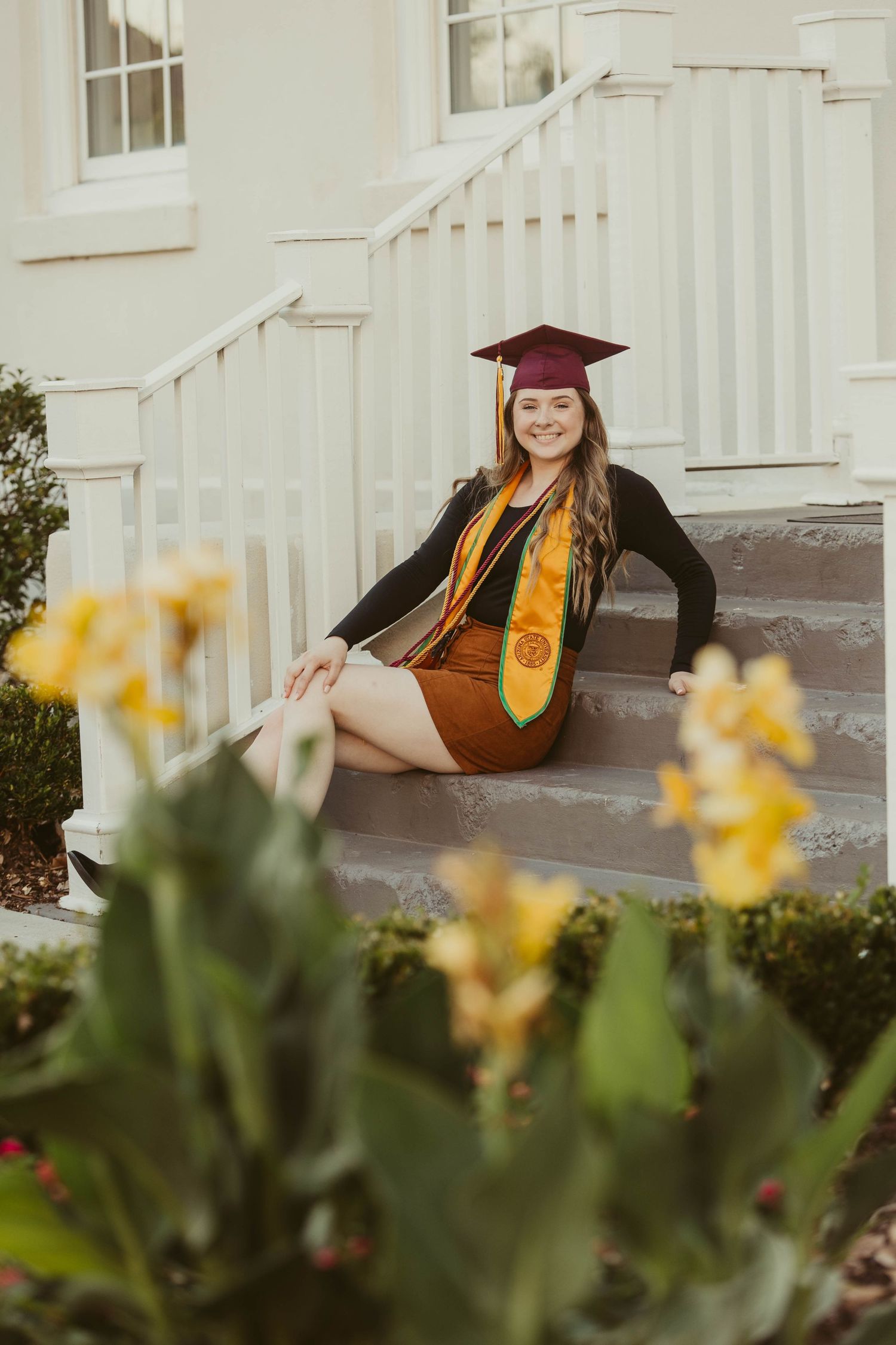 Graduate in academic regalia lounges on porch bench with yellow flowers in foreground.
