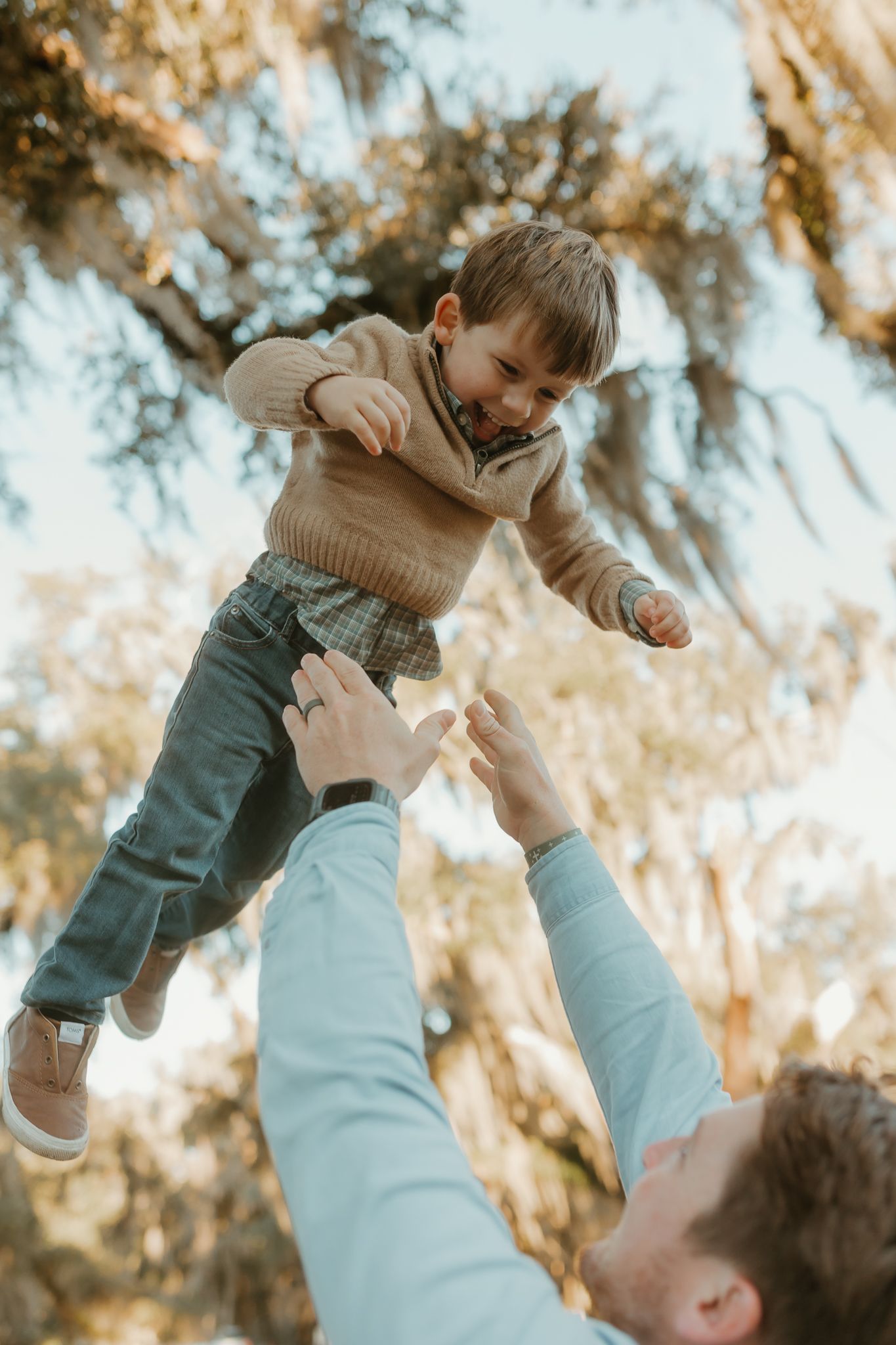 Playful moment of a child being lifted high into the air against a sunny forest backdrop.