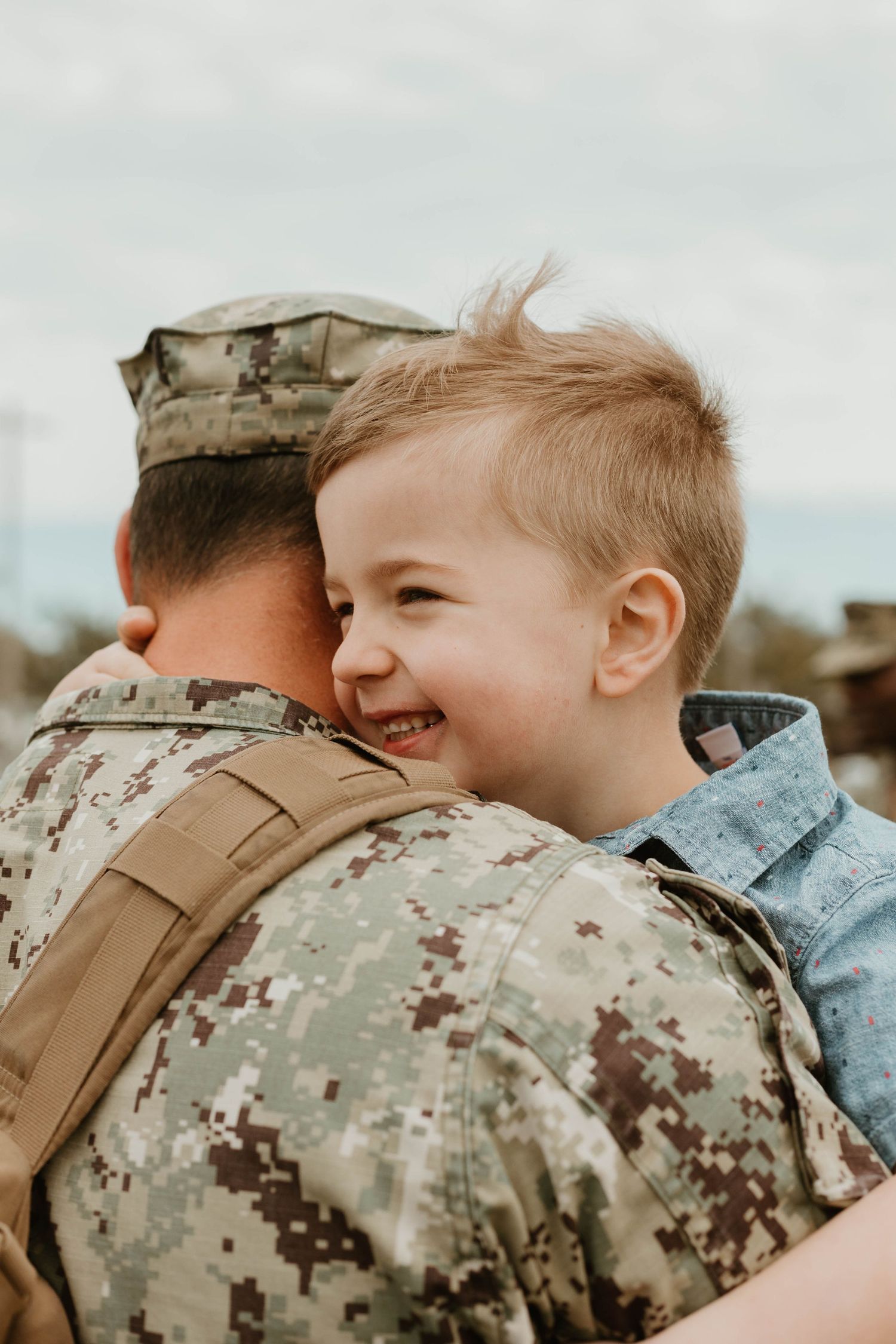 Young child smiling while hugging someone in military camouflage uniform.