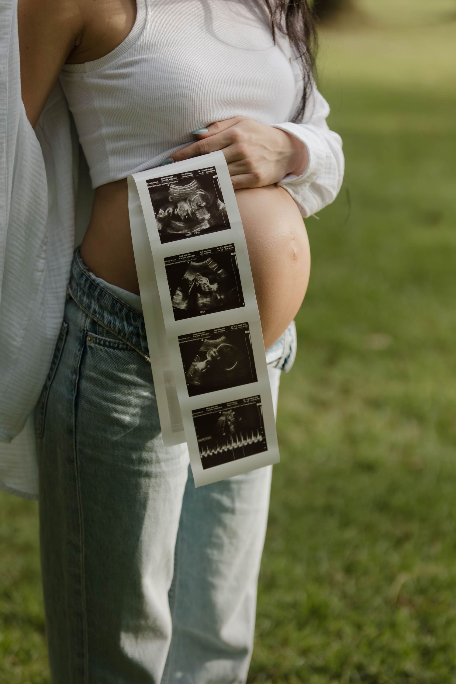 Expectant mother holding ultrasound photos against her pregnant belly outdoors.