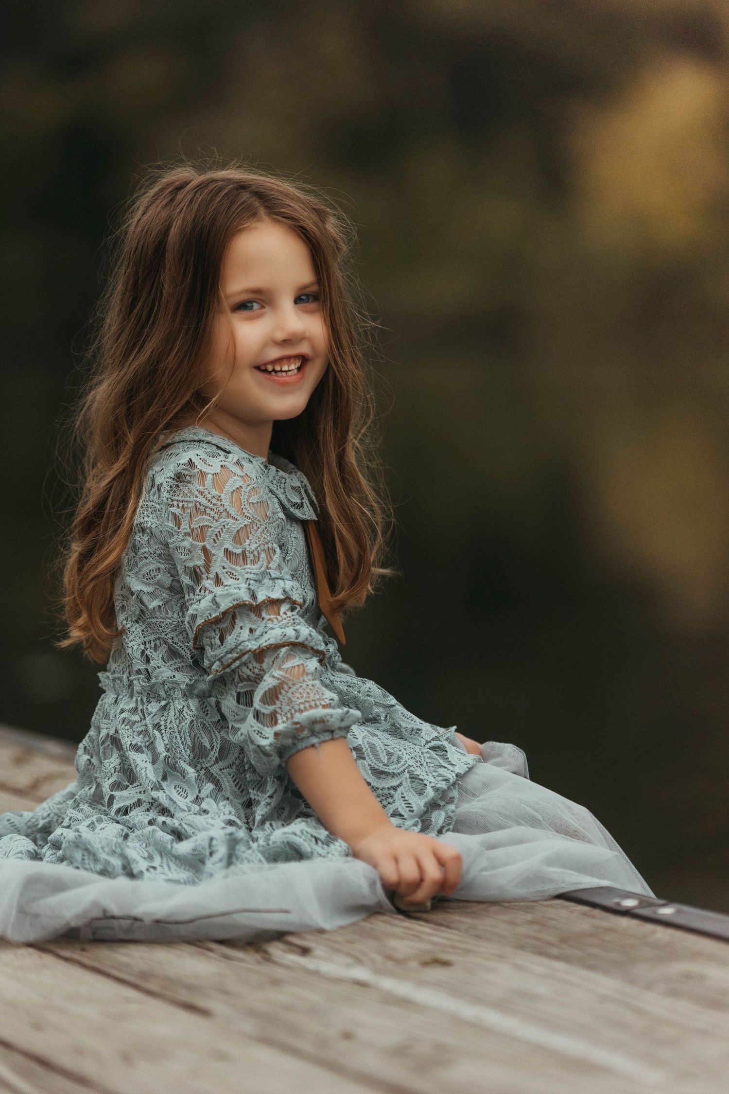 Child in blue lace dress poses playfully on wooden surface outdoors.