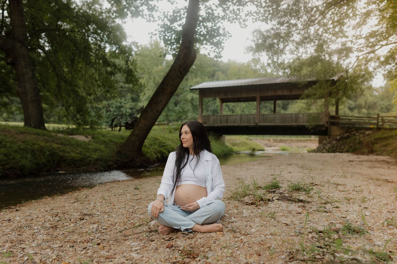 Maternity photo of an expectant mother sitting by a wooden bridge in a natural setting.
