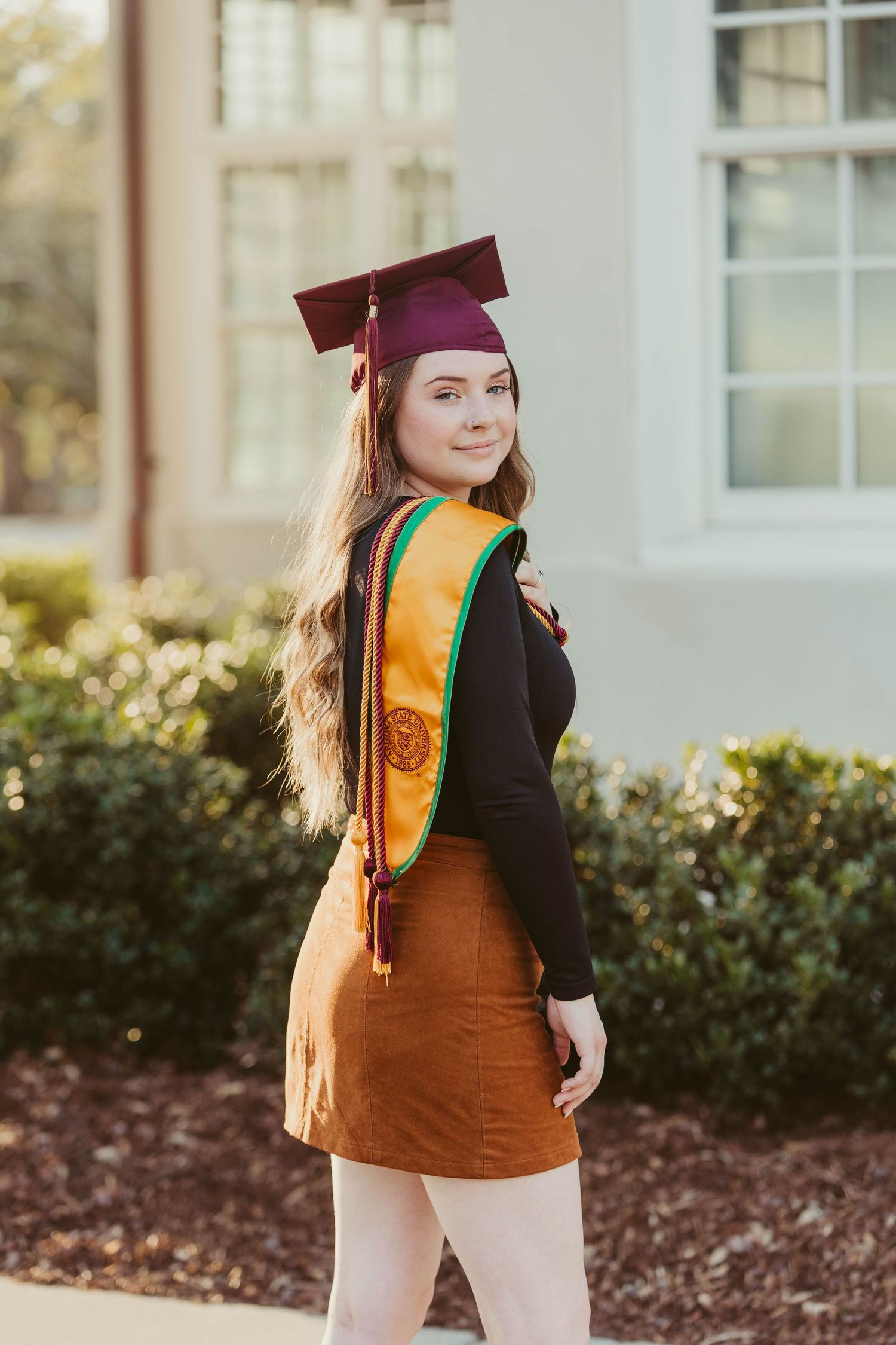 Graduate in burgundy cap and gold stole poses for outdoor graduation photos in front of white building.