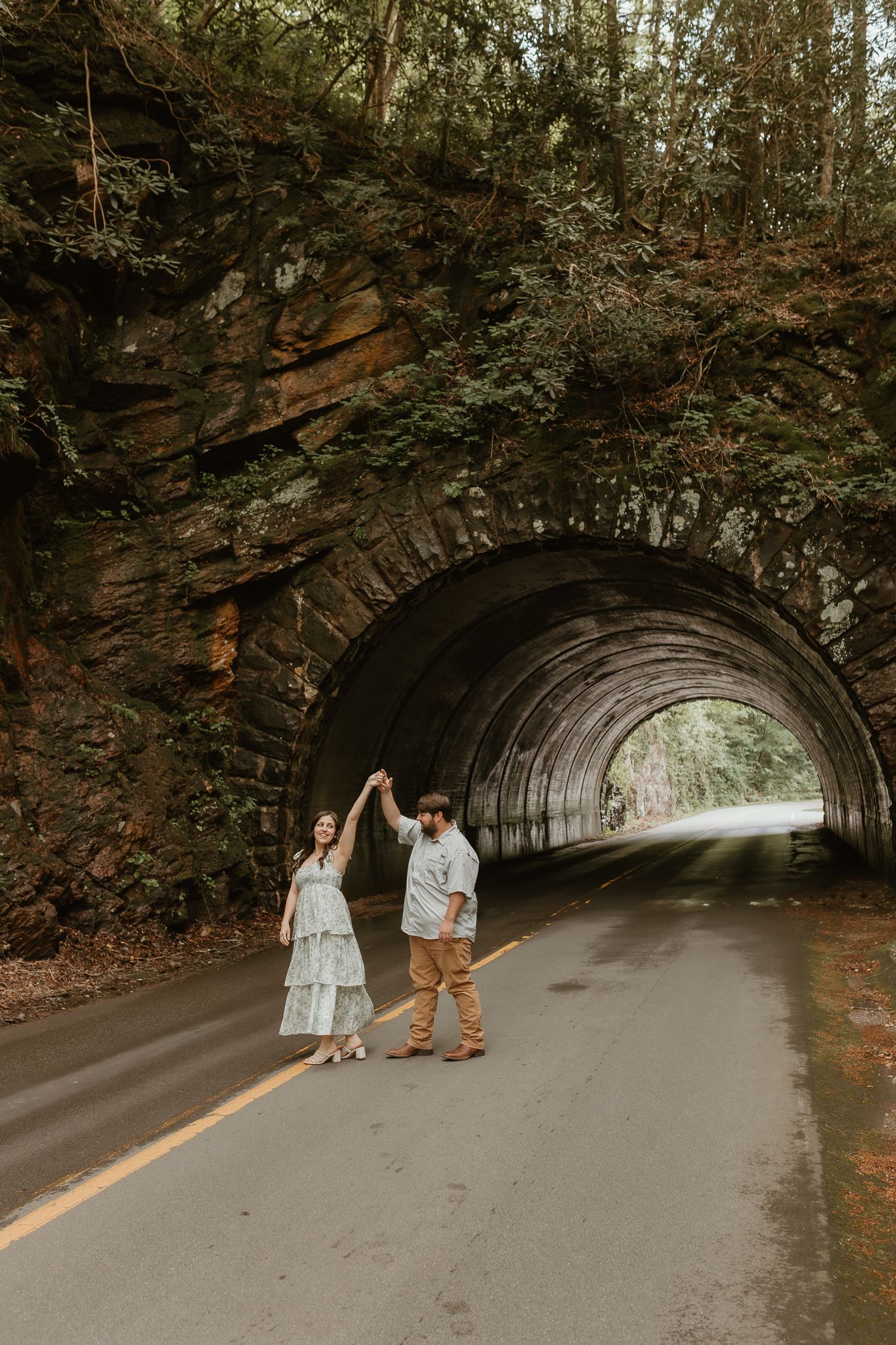 A couple strolls through a scenic stone tunnel on a paved road.