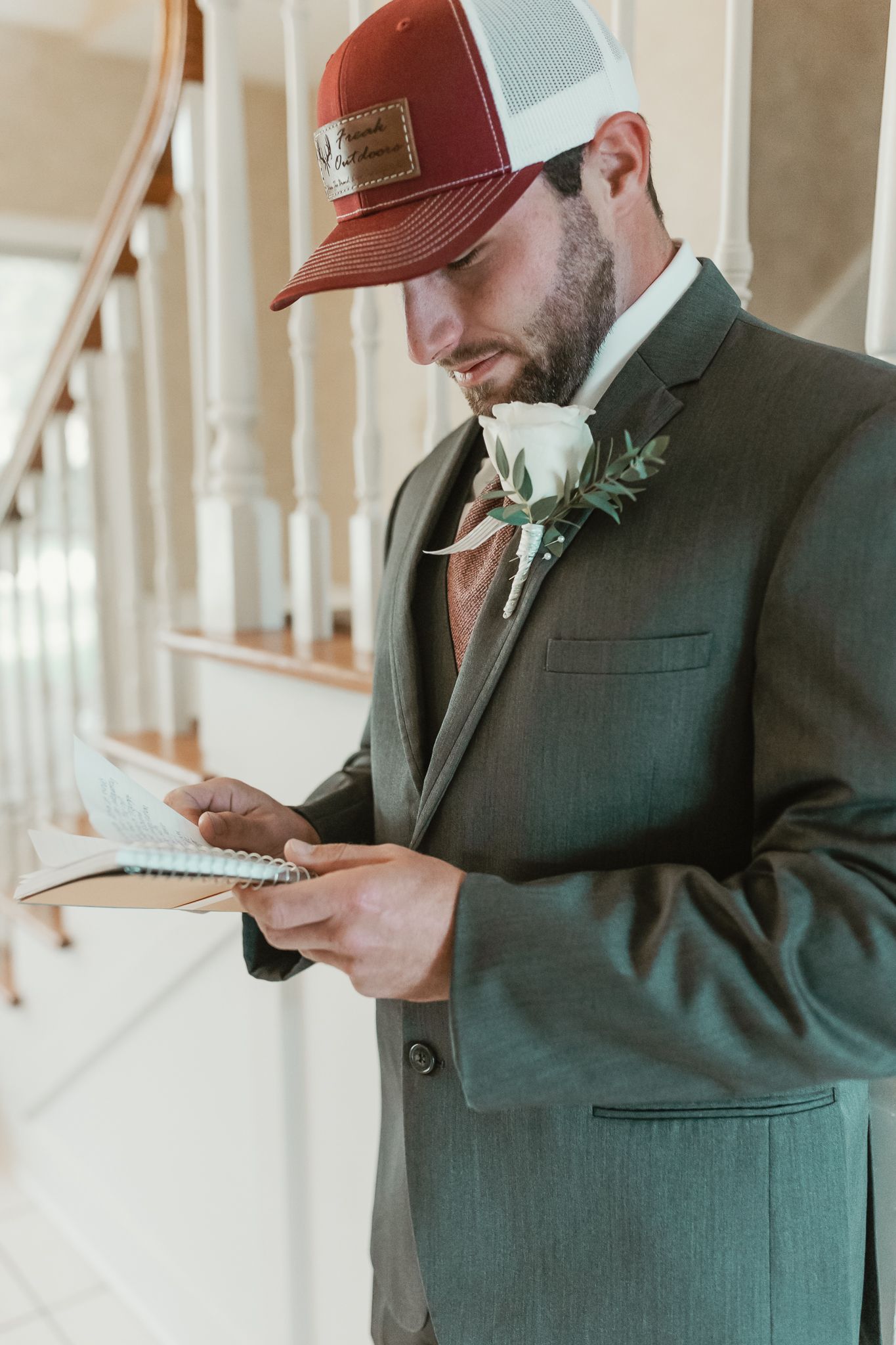 Groom in dark suit and baseball cap reading letter in sunlit hallway.