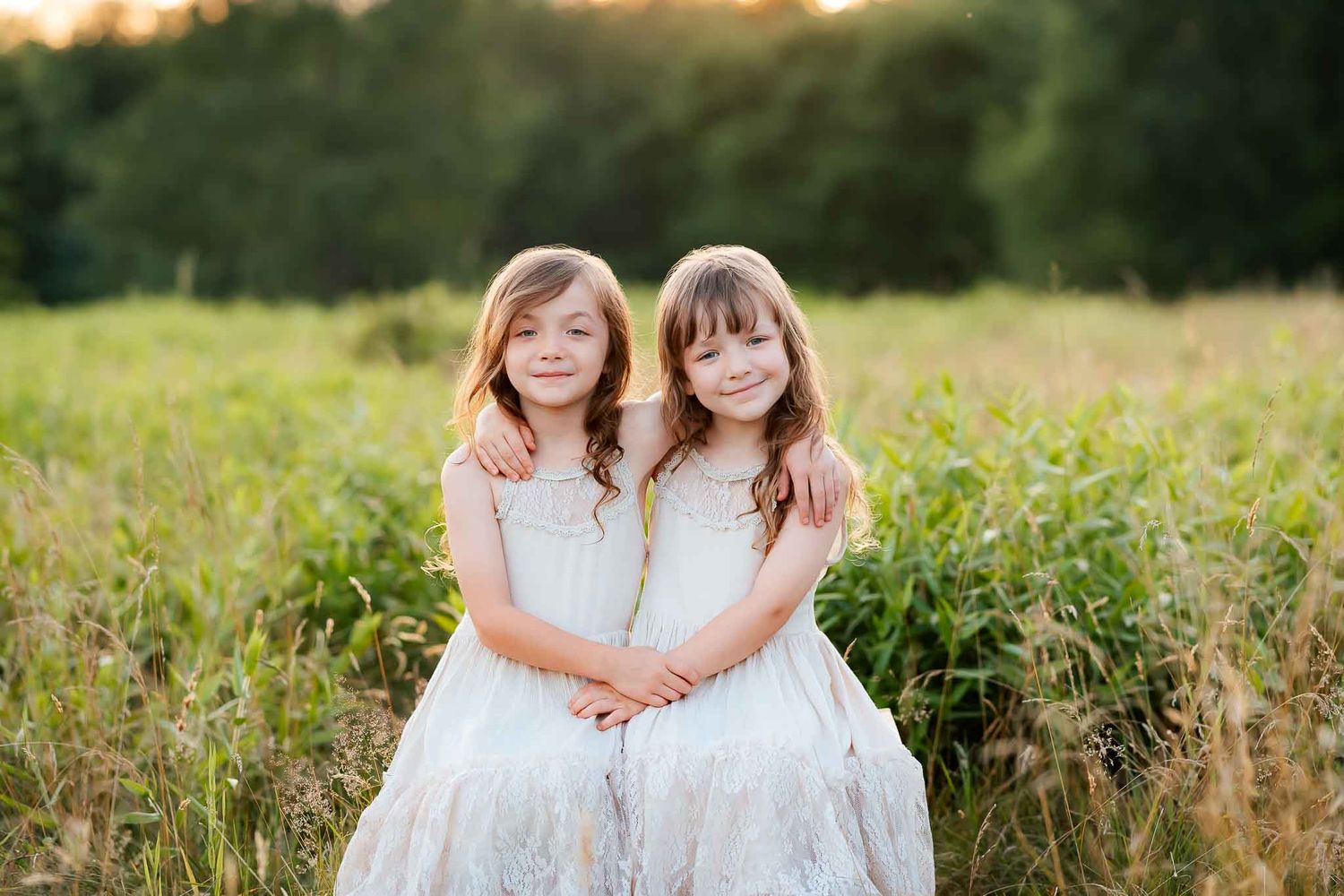 Two young girls in white dresses embrace while sitting in a sunlit meadow, smiling warmly for a portrait photo.