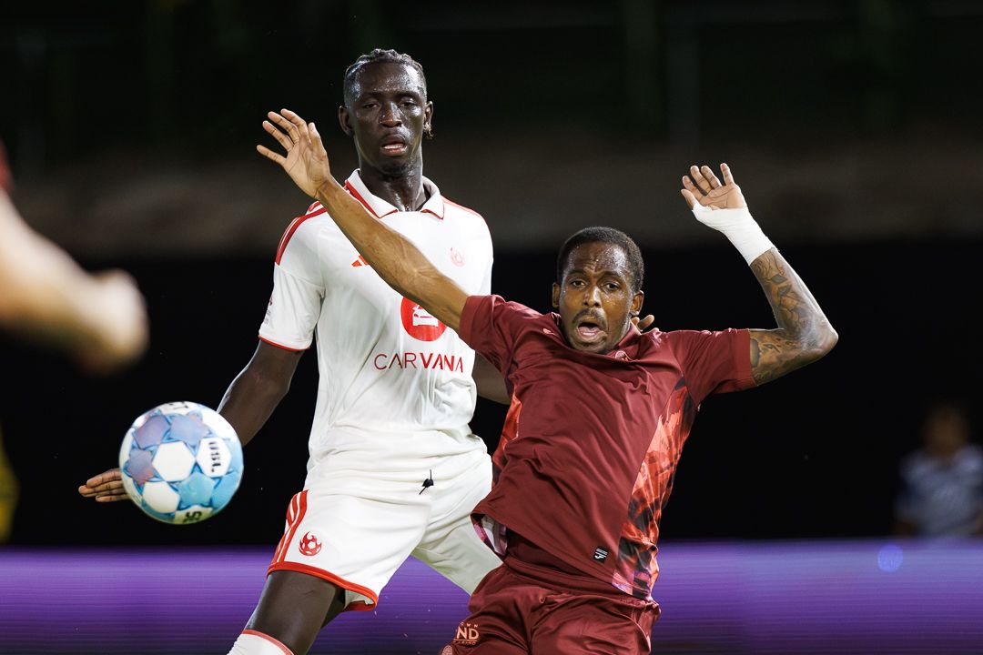 Soccer players battle for possession during a night match, one in white and one in maroon jersey.