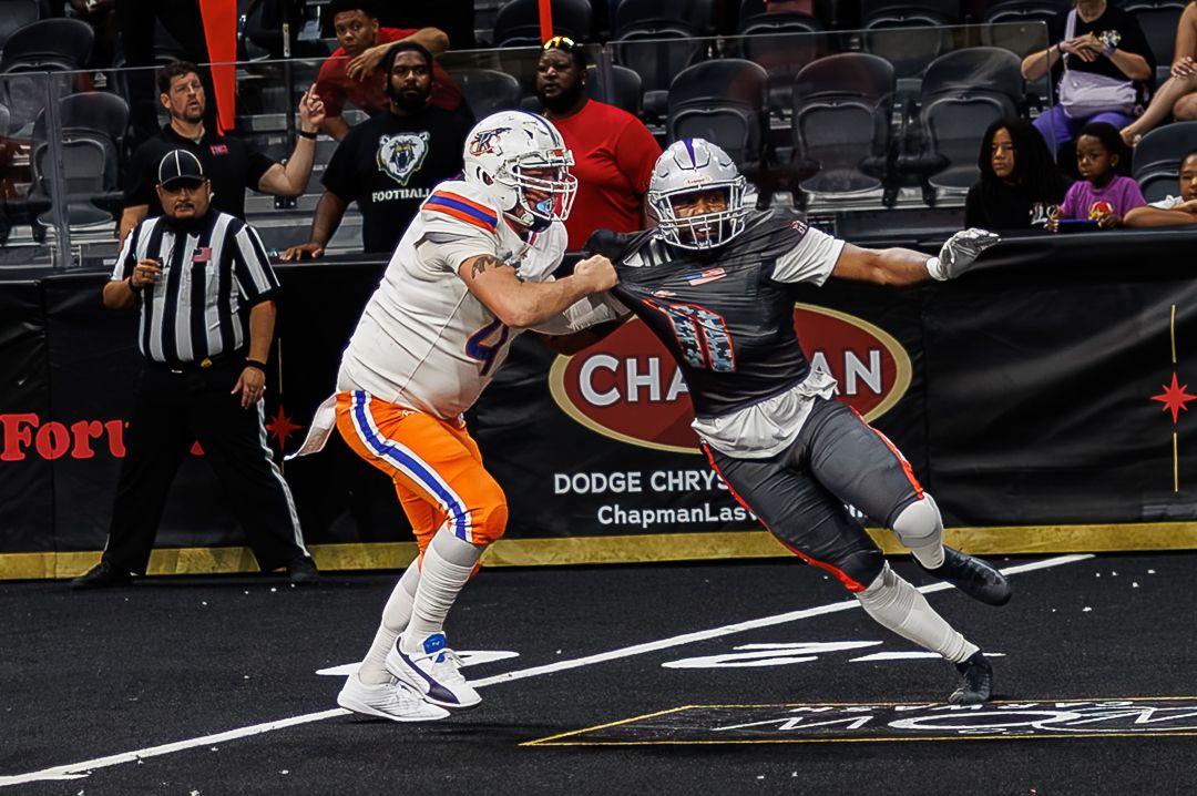 Two arena football players battle during an indoor game, with one in orange and white uniform blocking a gray-clad defender.