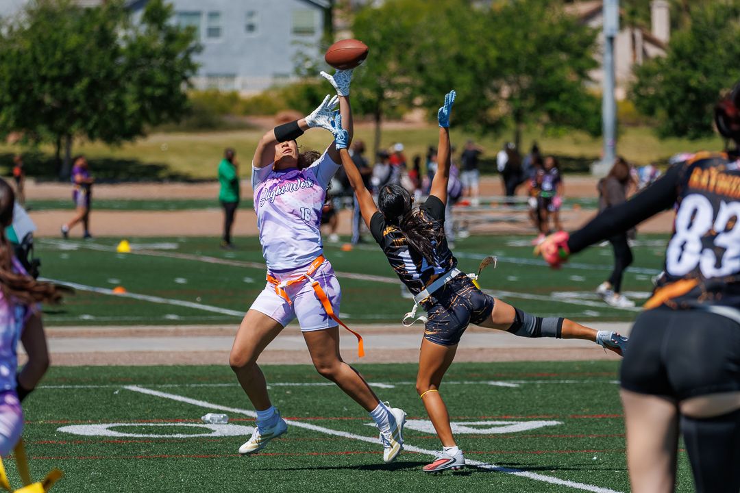 Players compete in a flag football game, with one athlete leaping to catch a pass under sunny skies on a green turf field.