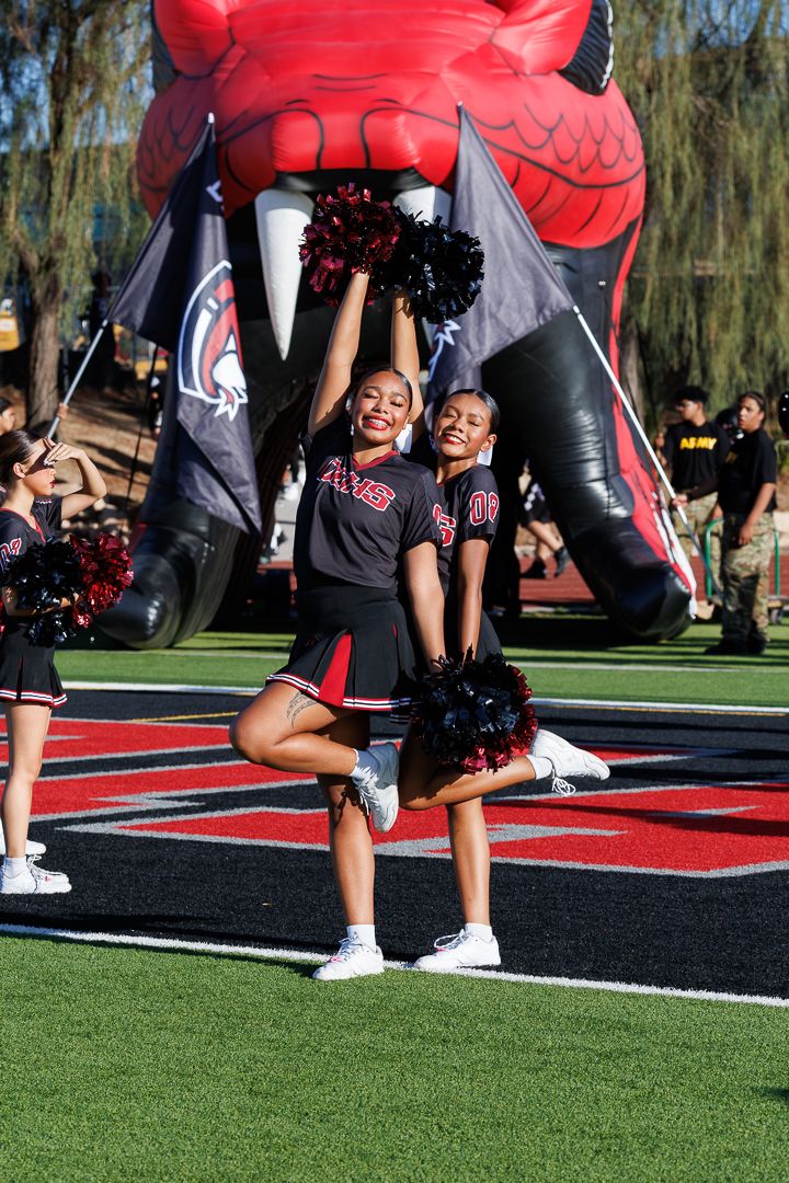 Two cheerleaders in black and red uniforms perform with pom-poms in front of an inflatable mascot at a football game.