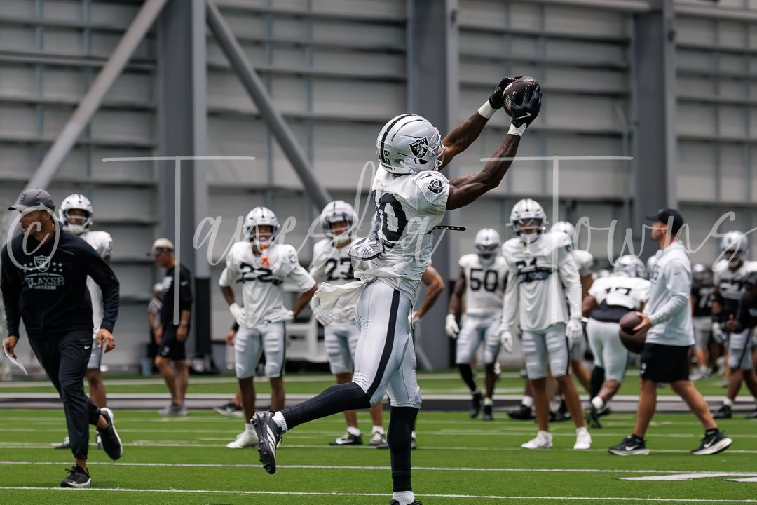 Football player in silver uniform leaping to catch a ball during an indoor practice session with teammates in background.