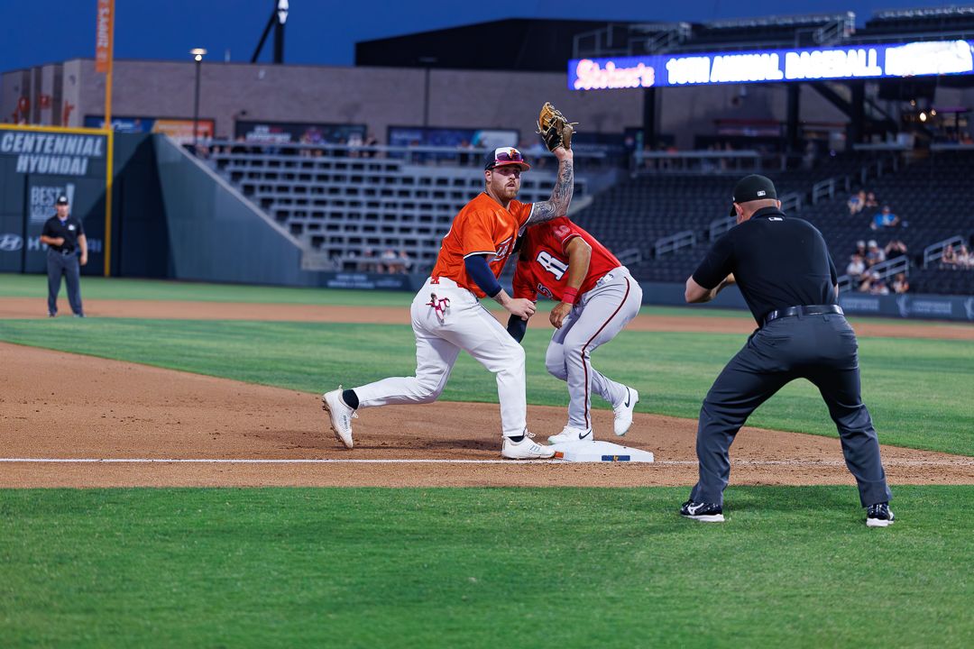 Baseball first baseman stretches to catch a throw as a runner approaches the bag during a night game.