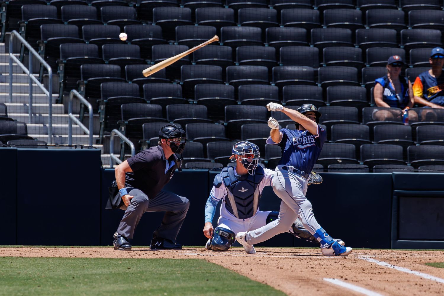 Baseball batter swings at pitch with catcher and umpire behind home plate in an empty stadium during a daytime game.