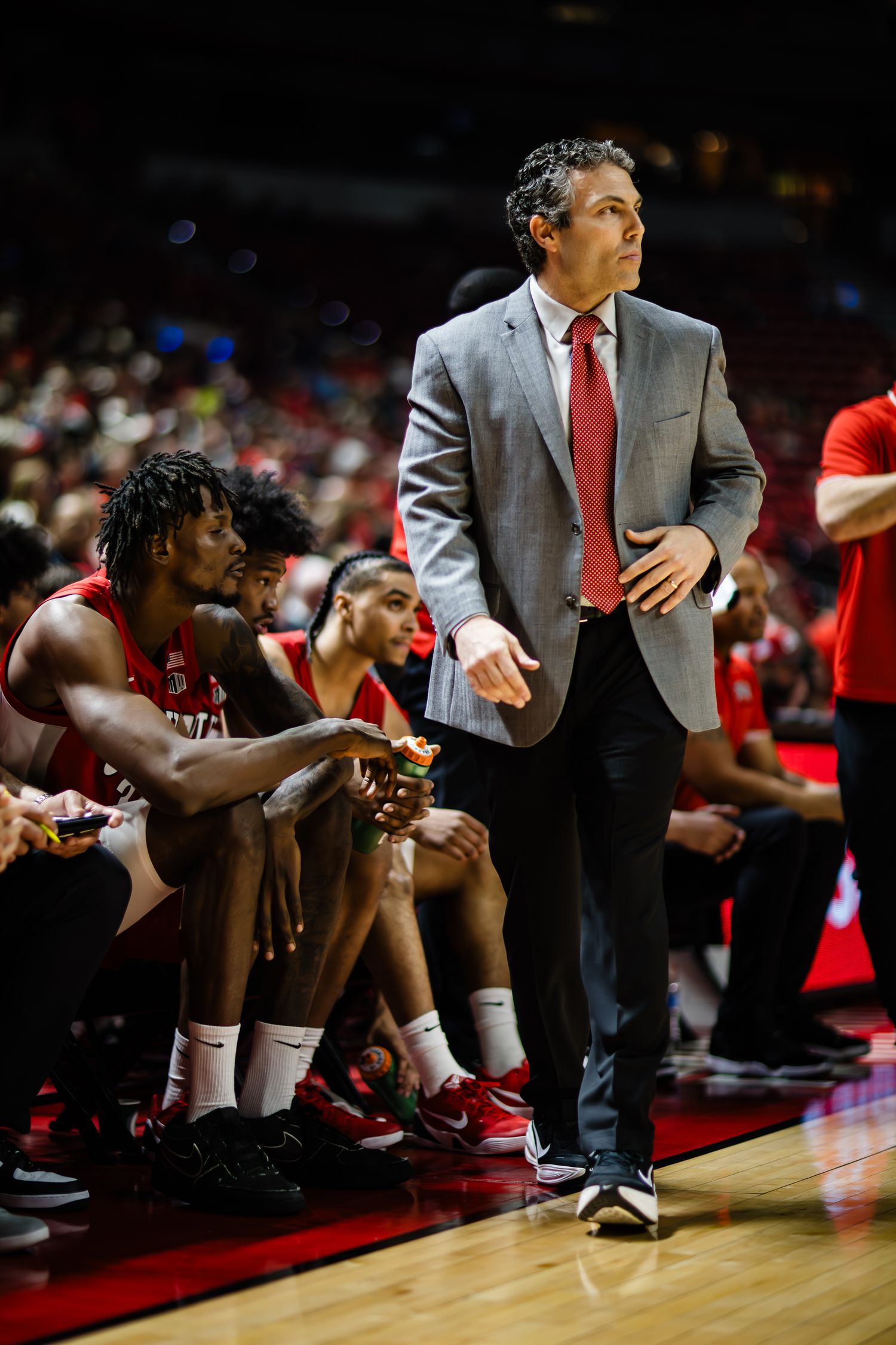 Basketball coach in gray suit gestures on sideline during game, players seated on bench behind him.
