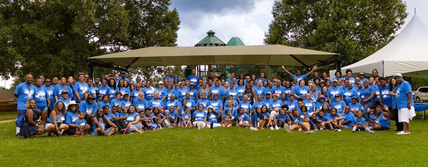 Large group photo of people in blue shirts sitting outdoors under trees.