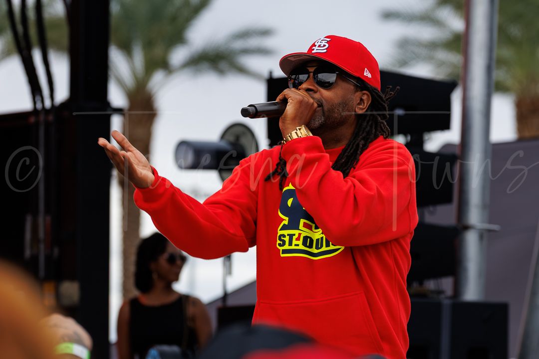 Rapper performing on outdoor stage wearing red St. Louis Cardinals hoodie and cap, singing into microphone.