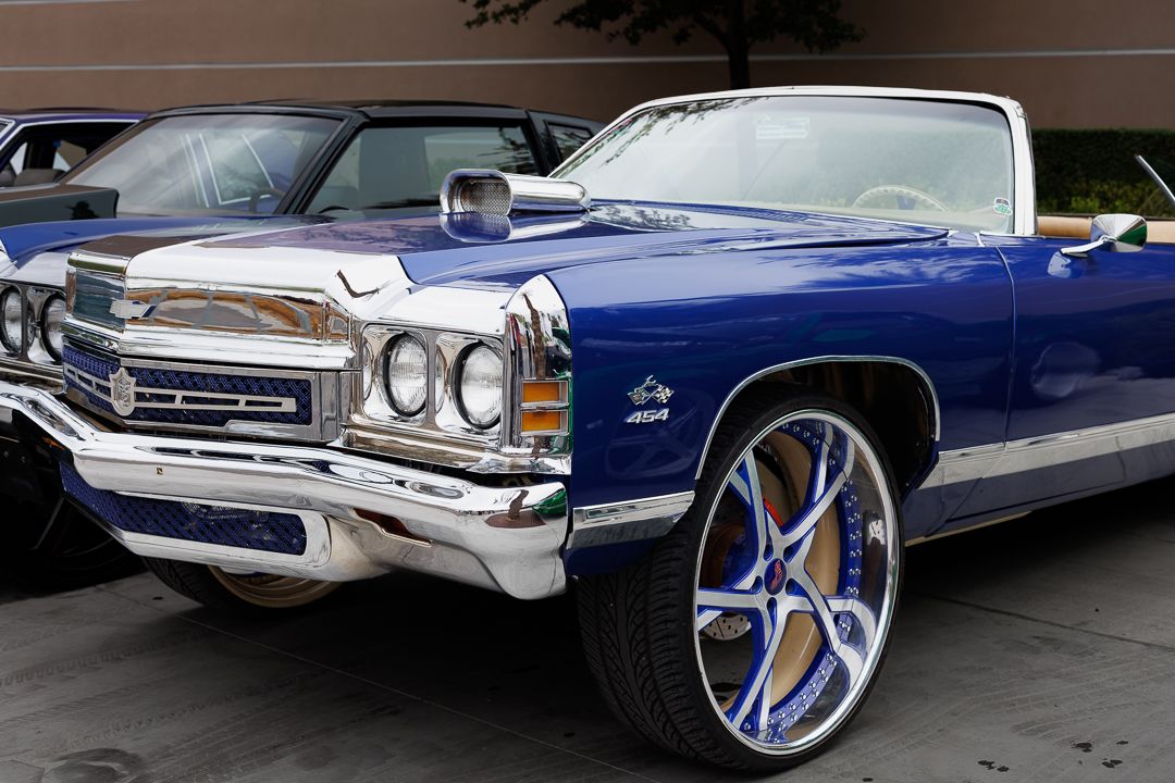 A classic blue Chevrolet Impala convertible with chrome accents and custom large white and blue rim wheels at a car show.