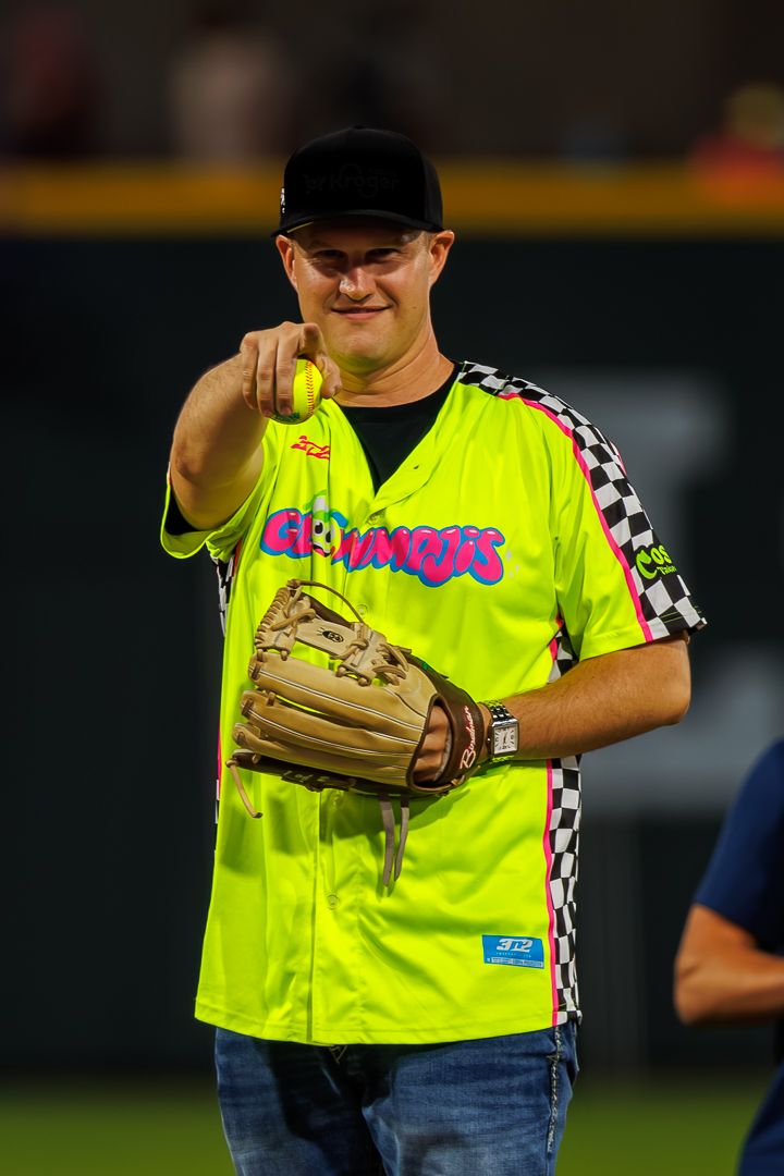 Man in neon yellow Growlers jersey holding baseball glove and pointing ball toward camera on baseball field.