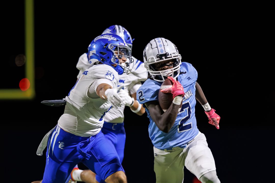 Two football players compete during a night game, one in blue and one in light blue jersey attempting a tackle.