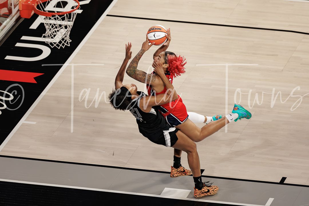 WNBA player in red uniform drives hard to the basket while defender attempts a block.