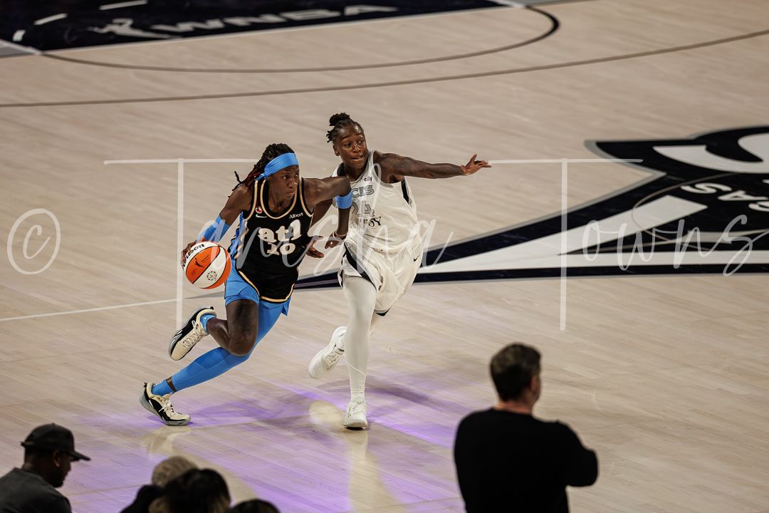 Two WNBA players compete during a basketball game, one dribbling while the other defends on a hardwood court.