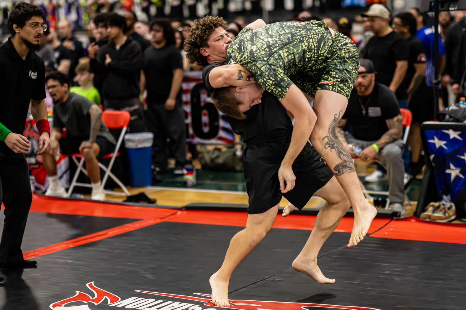 Two competitors grappling in a no-gi jiu-jitsu tournament, one athlete executing a takedown lift on a black mat.