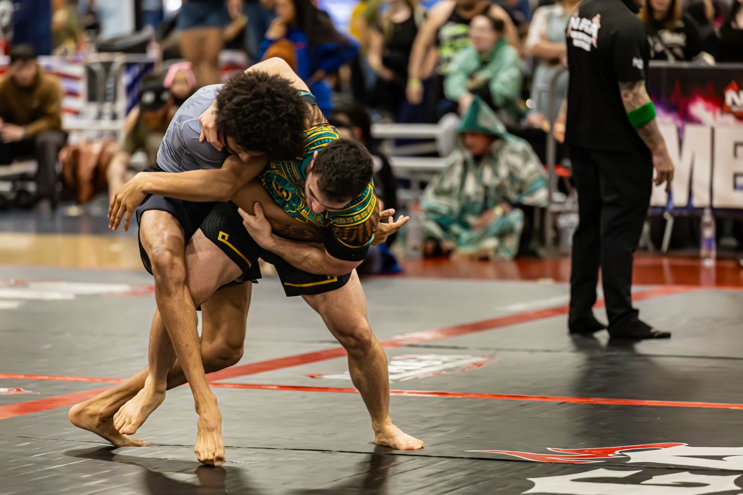 Two athletes compete in a grappling match on a mat at an indoor martial arts tournament.