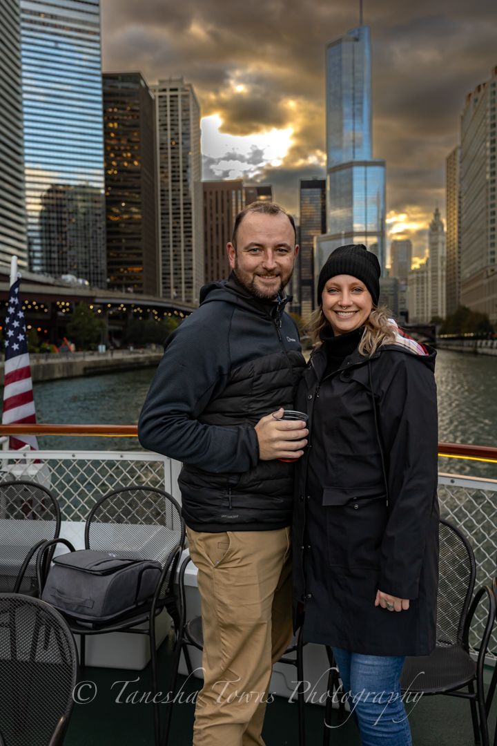 A couple poses for photos along Chicago's riverwalk at sunset with dramatic cloudy skies and city buildings in background.