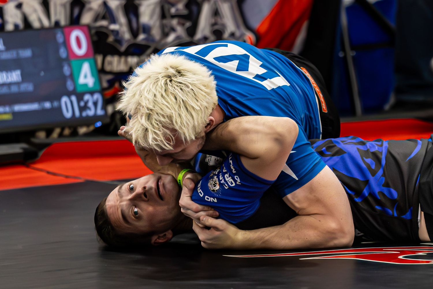 Two wrestlers compete on a mat, with one athlete in a blue uniform applying a submission hold during a grappling match.