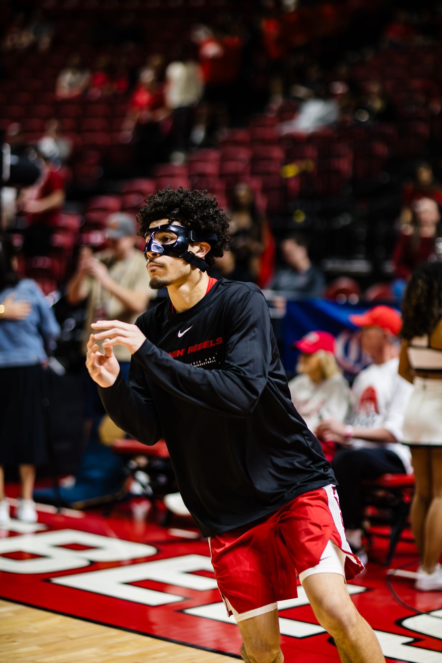 Basketball player wearing protective face mask and red shorts warming up on indoor court during game.
