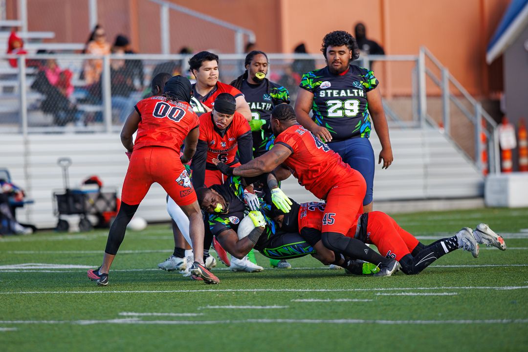 Football players in red and green uniforms compete in an intense play during a Stealth team game on a blue turf field.