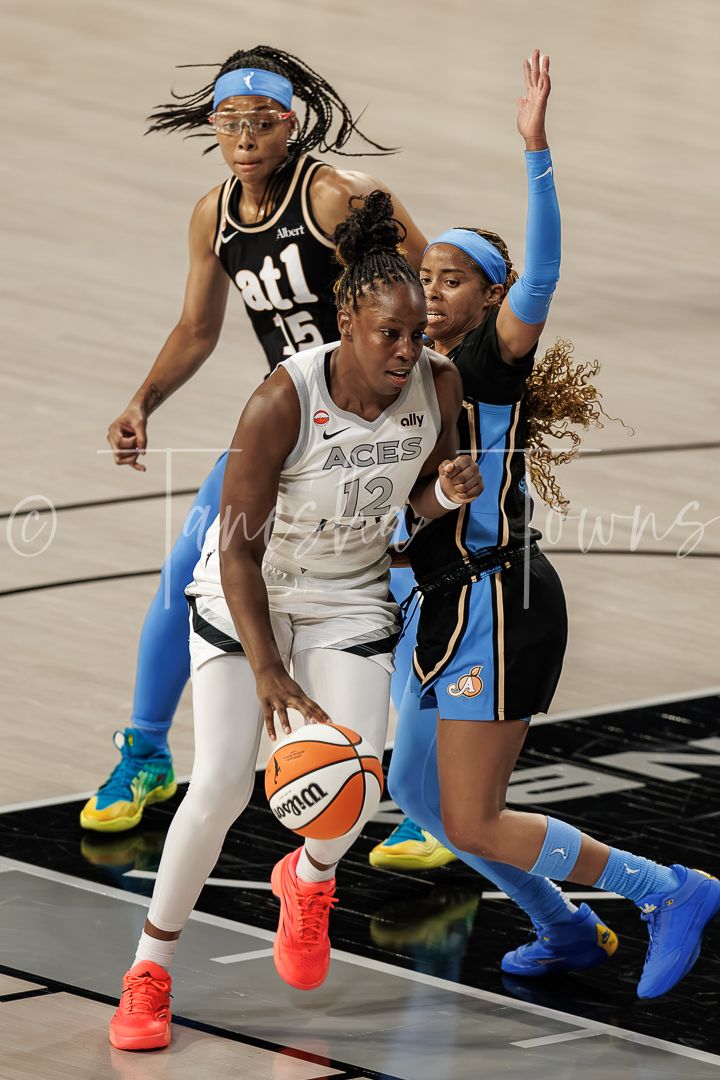 WNBA player #12 in white Aces uniform drives to basket against Chicago Sky defenders during professional game.