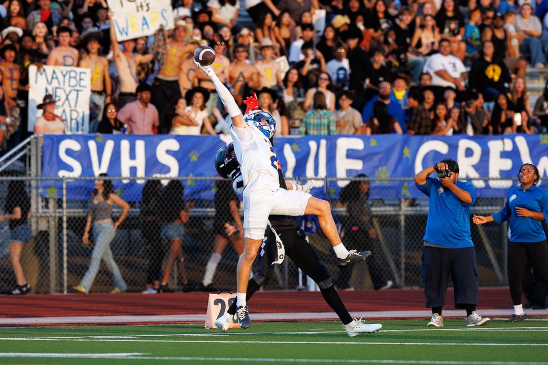 High school football player leaps to catch a pass during a night game at SVHS, with crowded bleachers in background.
