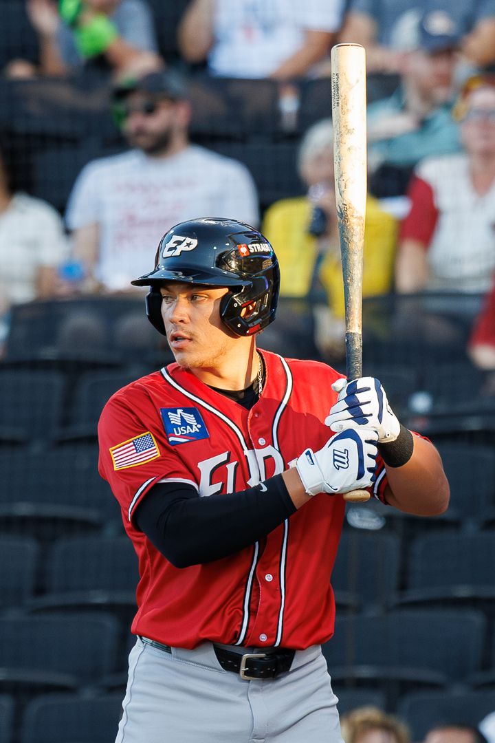 Baseball player in red EP uniform holds bat ready at plate, wearing helmet and batting gloves during a minor league game.